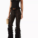 A woman wearing the Slip Top, a black lace slip top with adjustable shoulder straps, paired with matching sheer lace ruffle pants featuring peplum details. She is holding a small woven handbag and wearing black sandals.