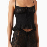 A woman wears the Slip Top—a black lace slip top with adjustable shoulder straps, a tied detail at the bust, and a slightly ruffled hem—paired with matching black lace pants featuring a tie at the waist.