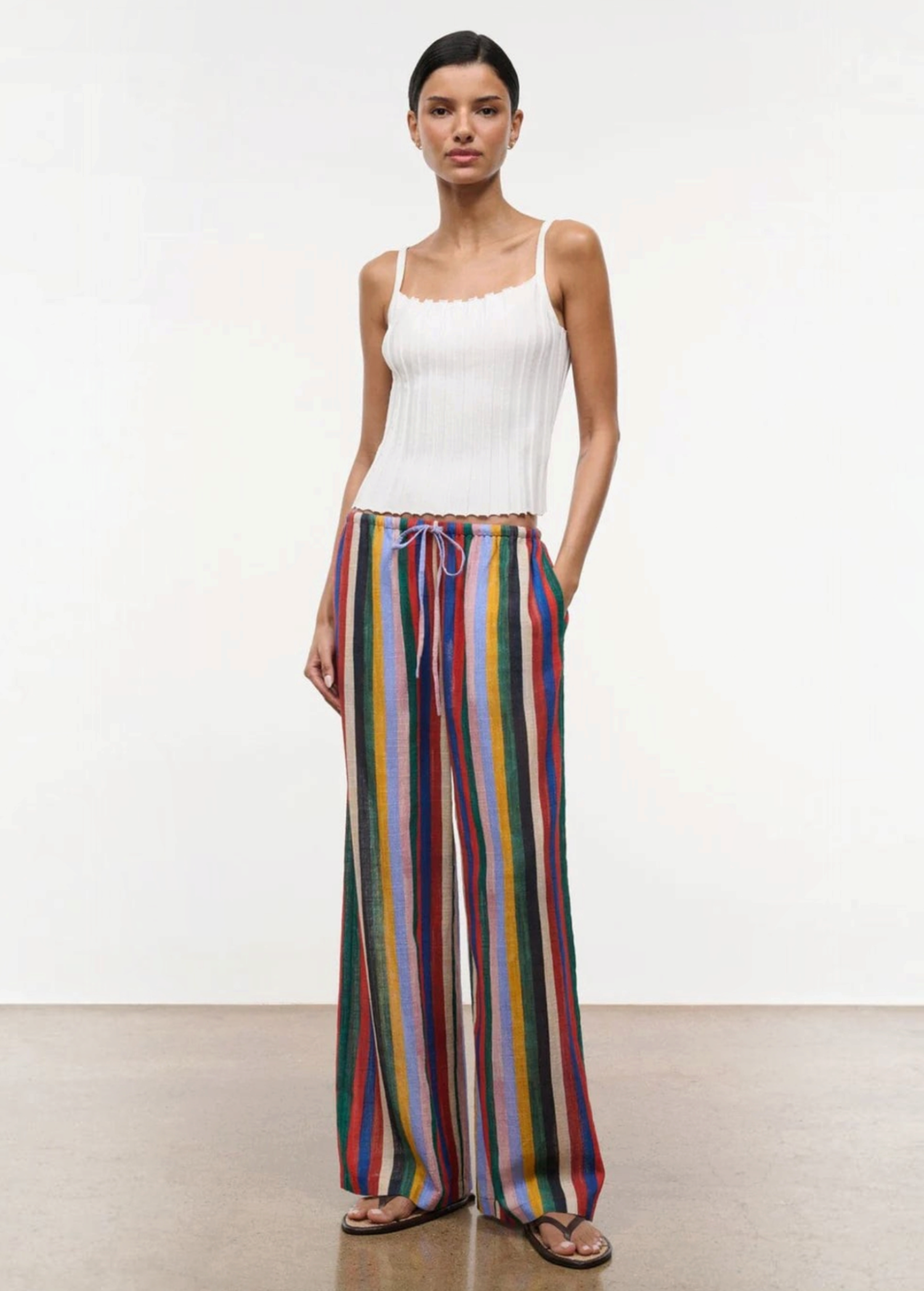 A model wears a white ribbed tank top with the Mateo Pant, which are relaxed high-waisted, wide-leg pants featuring vertical multicolored stripes and a drawstring waist.
