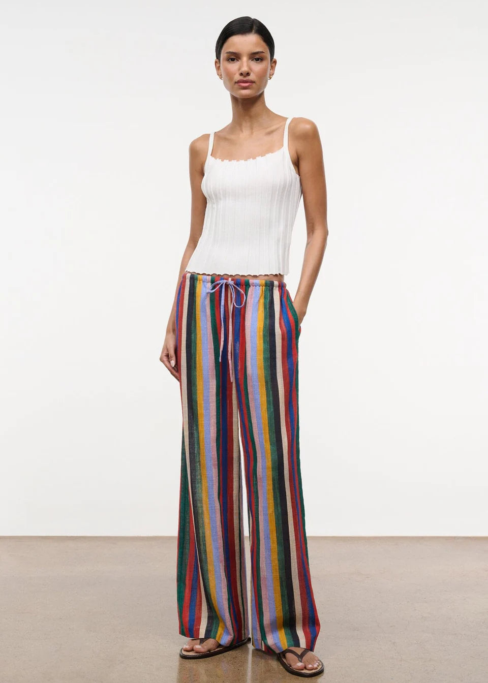 A model wears a white ribbed tank top with the Mateo Pant, which are relaxed high-waisted, wide-leg pants featuring vertical multicolored stripes and a drawstring waist.