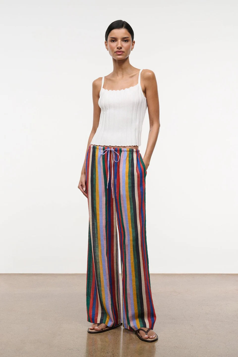 A model wears a white ribbed tank top with the Mateo Pant, which are relaxed high-waisted, wide-leg pants featuring vertical multicolored stripes and a drawstring waist.