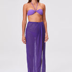 A woman is shown barefoot on a plain white background, wearing the Sarong Skirt in sheer pleated fabric with matching purple tones and a bandeau bikini top. She looks slightly to her left with a neutral expression.