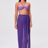 A woman is shown barefoot on a plain white background, wearing the Sarong Skirt in sheer pleated fabric with matching purple tones and a bandeau bikini top. She looks slightly to her left with a neutral expression.