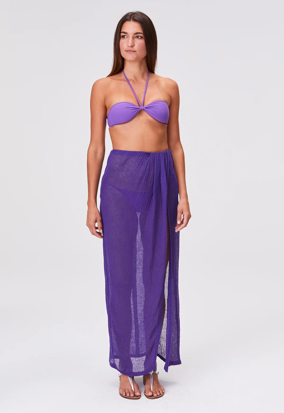 A woman is shown barefoot on a plain white background, wearing the Sarong Skirt in sheer pleated fabric with matching purple tones and a bandeau bikini top. She looks slightly to her left with a neutral expression.