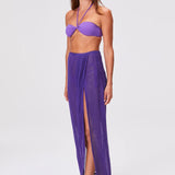 A woman models the Sarong Skirt—a sheer, purple pleated design with a high slit—styled with a coordinating purple bandeau bikini top and clear sandals, set against a plain light gray background.