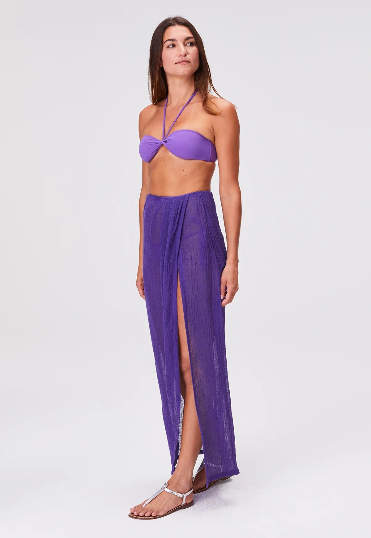 A woman models the Sarong Skirt—a sheer, purple pleated design with a high slit—styled with a coordinating purple bandeau bikini top and clear sandals, set against a plain light gray background.