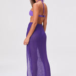 A woman stands sideways wearing a purple halter bikini top, the Sarong Skirt—a sheer, floor-length pleated skirt—and sandals, set against a plain white background.