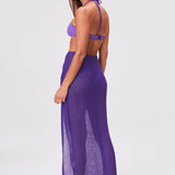 A woman stands sideways wearing a purple halter bikini top, the Sarong Skirt—a sheer, floor-length pleated skirt—and sandals, set against a plain white background.