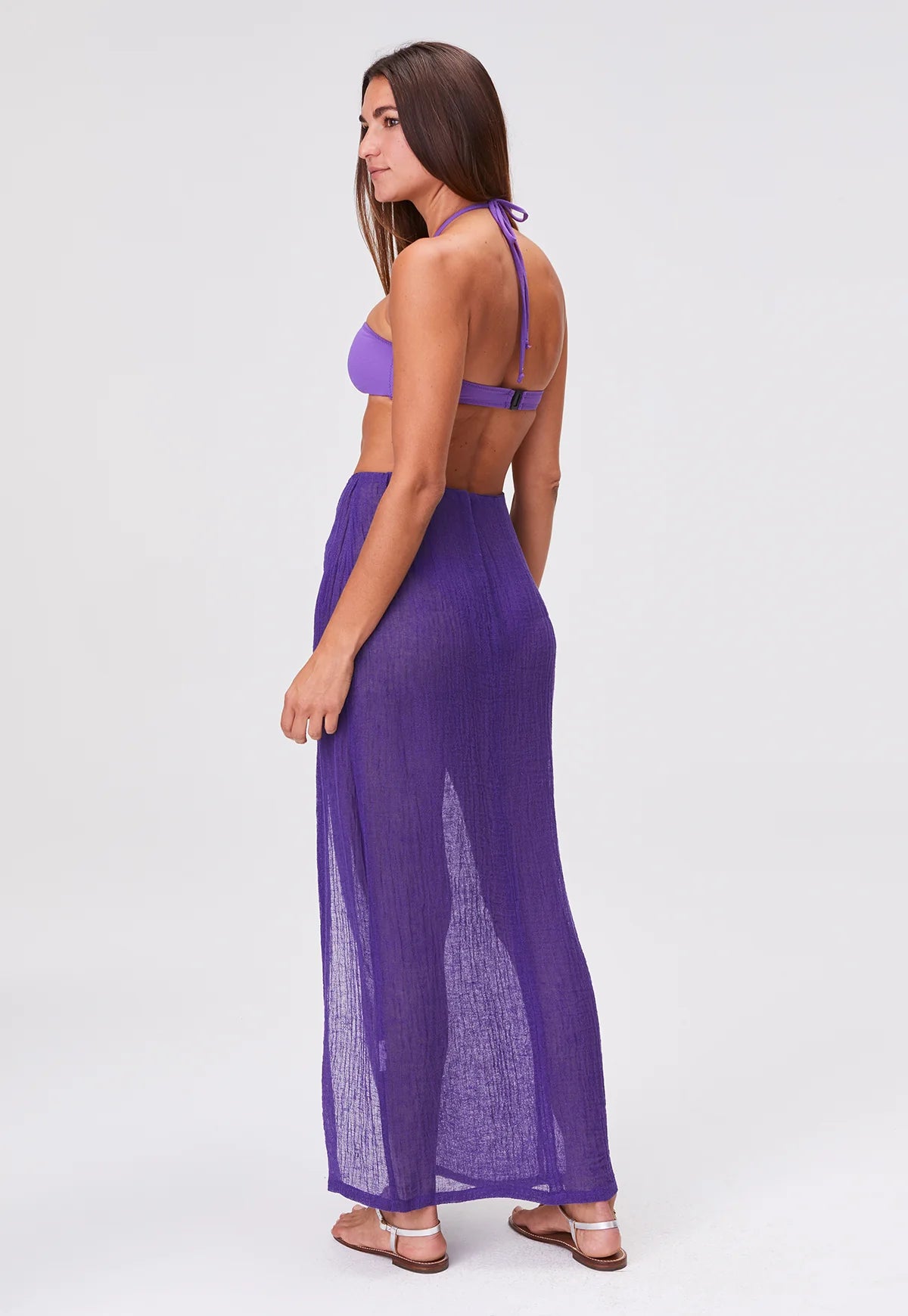 A woman stands sideways wearing a purple halter bikini top, the Sarong Skirt—a sheer, floor-length pleated skirt—and sandals, set against a plain white background.