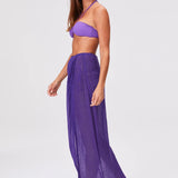 A woman wearing a purple bikini top, sheer Sarong Skirt, and sandals stands in profile against a plain white background.