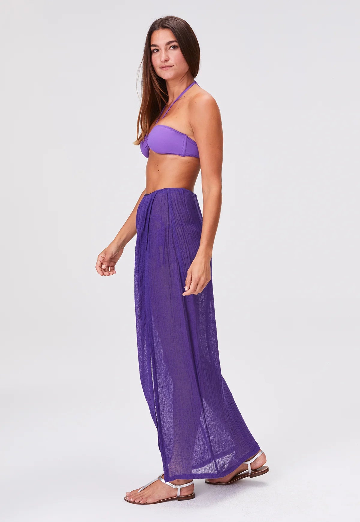 A woman wearing a purple bikini top, sheer Sarong Skirt, and sandals stands in profile against a plain white background.