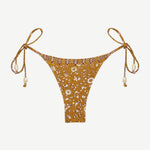 Praia Bottom: A gold beaded bikini bottom featuring a delicate white floral print, thin adjustable side ties embellished with beads, and a high-cut silhouette, made from recycled nylon swimwear material and showcased on a plain white background.