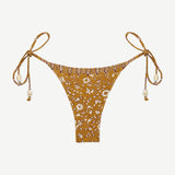 Praia Bottom: A gold beaded bikini bottom featuring a delicate white floral print, thin adjustable side ties embellished with beads, and a high-cut silhouette, made from recycled nylon swimwear material and showcased on a plain white background.
