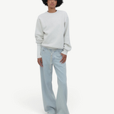 Person with curly hair wearing a white sweatshirt, Cool Jeans in light blue made from the softest denim, and white sneakers stands against a plain, light gray background.