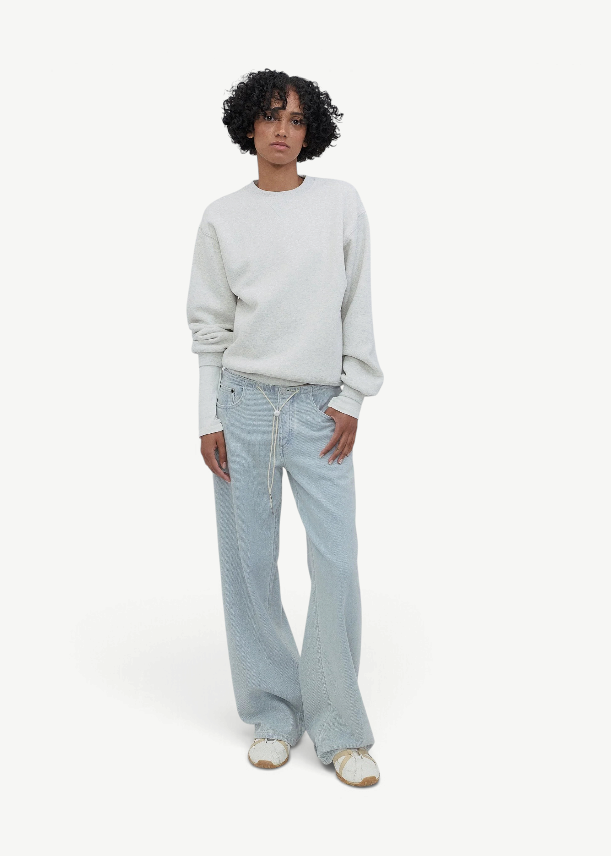 Person with curly hair wearing a white sweatshirt, Cool Jeans in light blue made from the softest denim, and white sneakers stands against a plain, light gray background.
