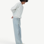 A person with curly hair wearing a white sweater, Cool Jeans in a light blue relaxed leg fit crafted from the softest denim, and white shoes stands in a minimalist setting with hands behind their back.