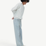 A person with curly hair wearing a white sweater, Cool Jeans in a light blue relaxed leg fit crafted from the softest denim, and white shoes stands in a minimalist setting with hands behind their back.