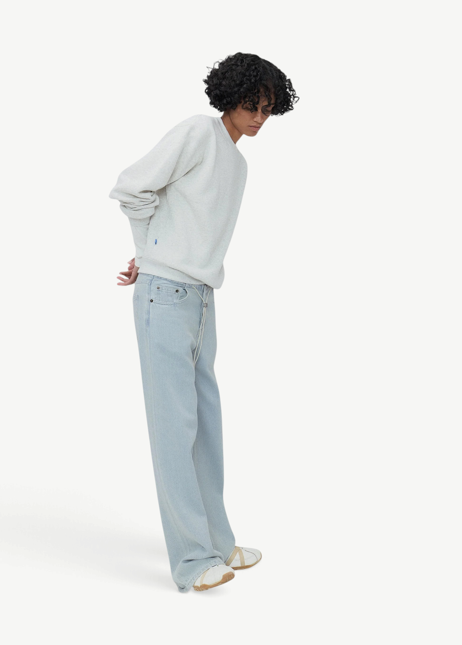 A person with curly hair wearing a white sweater, Cool Jeans in a light blue relaxed leg fit crafted from the softest denim, and white shoes stands in a minimalist setting with hands behind their back.
