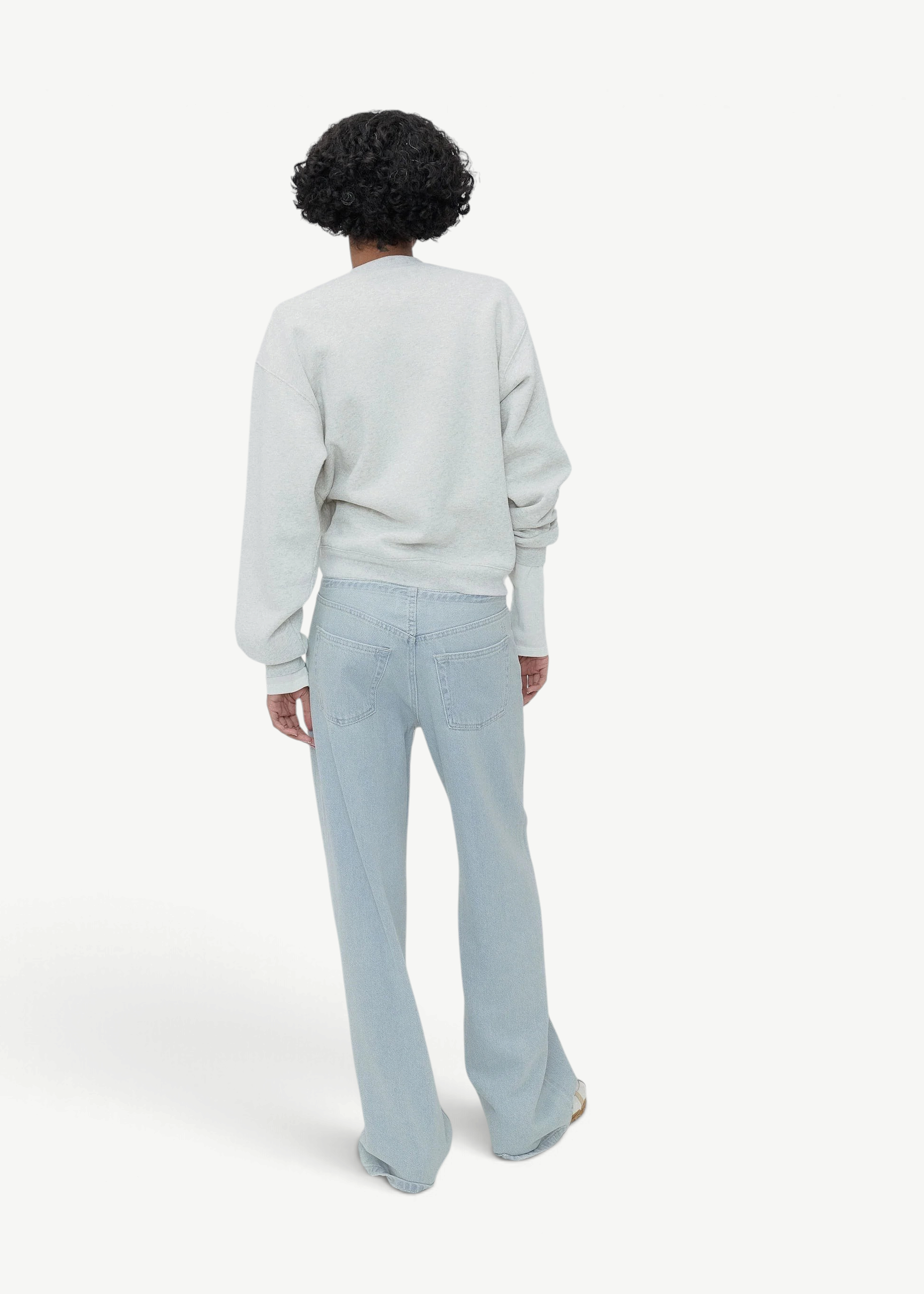 Wearing Cool Jeans in the softest denim, a person with short curly hair stands facing away, paired with a light grey sweatshirt against a plain, light grey background.