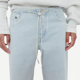 A model wears Cool Jeans—light wash, relaxed leg jeans secured with a white drawstring in place of a belt or button—paired with a light grey sweatshirt. The image features the torso and upper legs, with hands resting naturally at the sides.