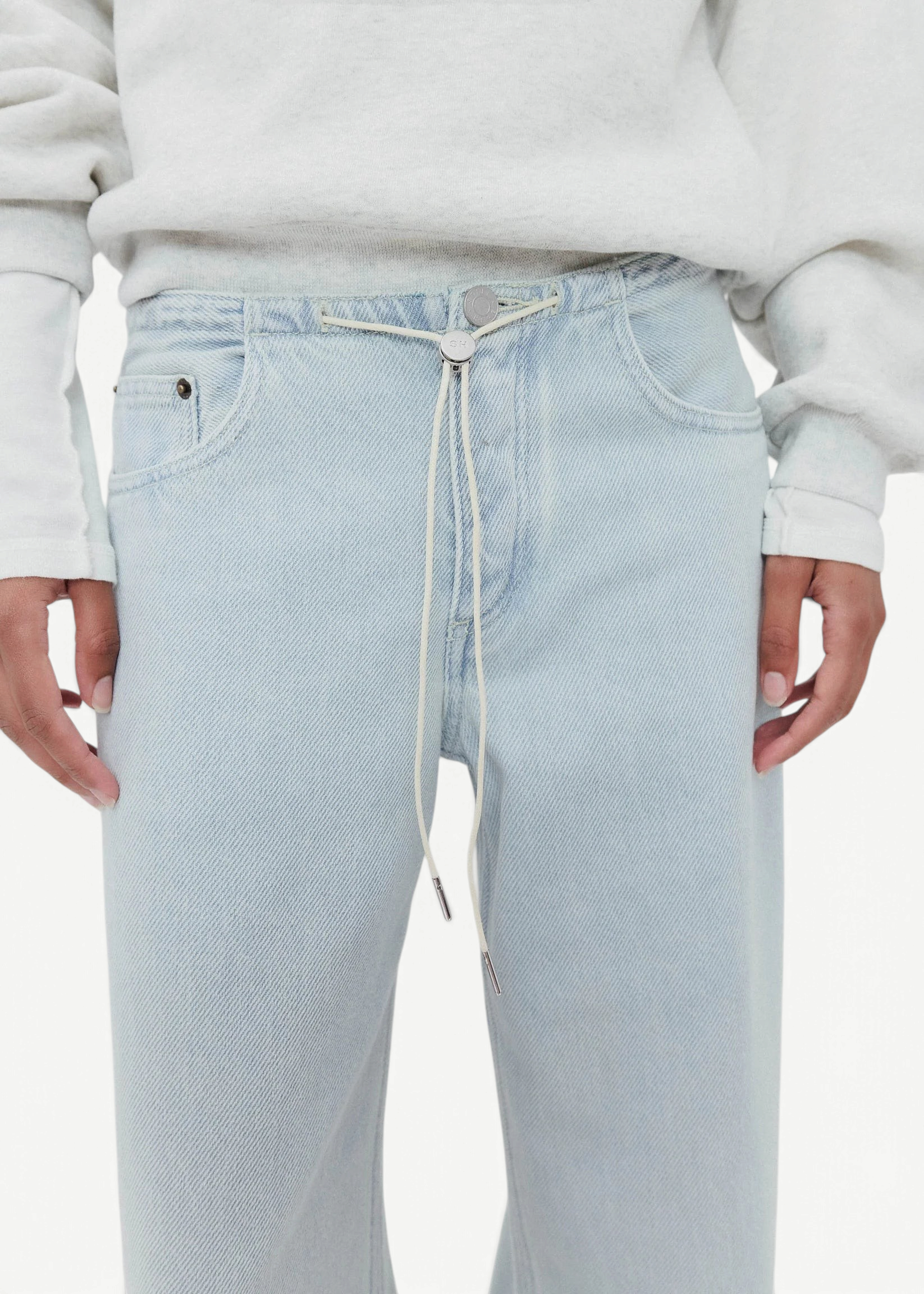 A model wears Cool Jeans—light wash, relaxed leg jeans secured with a white drawstring in place of a belt or button—paired with a light grey sweatshirt. The image features the torso and upper legs, with hands resting naturally at the sides.
