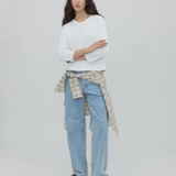 A woman with long dark hair stands against a gray background, wearing a white top, black shoes, and light blue Everyday Jeans featuring a relaxed silhouette, with a plaid shirt tied around her waist.