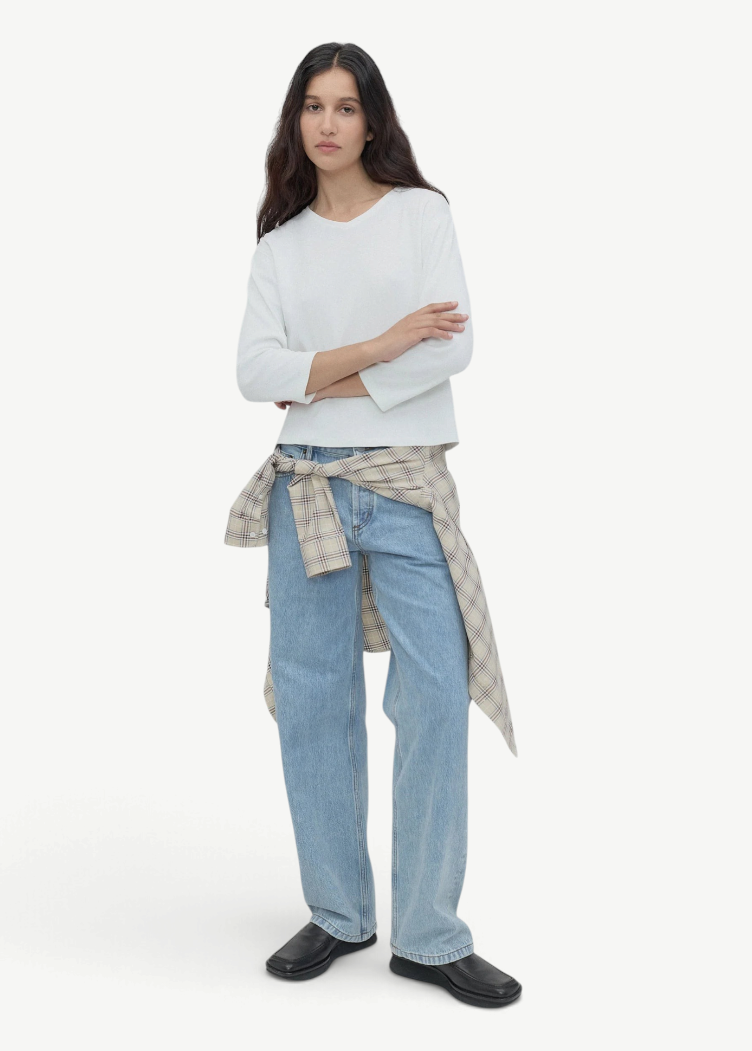 A woman with long dark hair stands against a gray background, wearing a white top, black shoes, and light blue Everyday Jeans featuring a relaxed silhouette, with a plaid shirt tied around her waist.