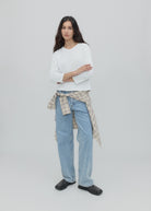 A woman with long dark hair stands against a gray background, wearing a white top, black shoes, and light blue Everyday Jeans featuring a relaxed silhouette, with a plaid shirt tied around her waist.
