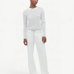 A person stands against a plain white background, wearing a white long-sleeved shirt with a collar, paired with the Cool Jean—white relaxed leg jeans featuring an adjustable drawstring—and beige shoes. They have short hair and a relaxed expression, with arms resting by their sides.