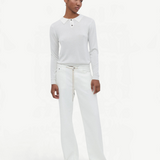 A person stands against a plain white background, wearing a white long-sleeved shirt with a collar, paired with the Cool Jean—white relaxed leg jeans featuring an adjustable drawstring—and beige shoes. They have short hair and a relaxed expression, with arms resting by their sides.