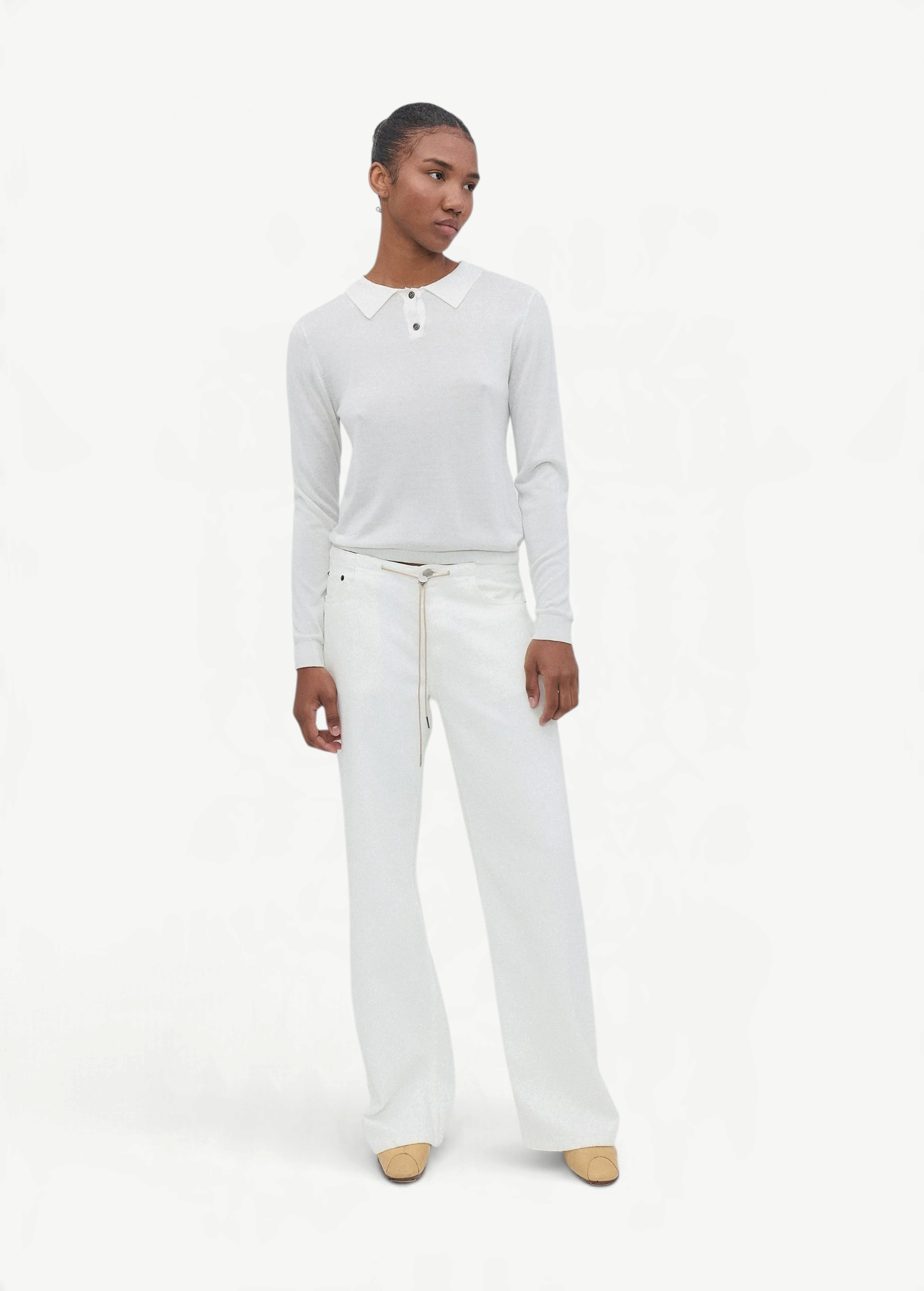 A person stands against a plain white background, wearing a white long-sleeved shirt with a collar, paired with the Cool Jean—white relaxed leg jeans featuring an adjustable drawstring—and beige shoes. They have short hair and a relaxed expression, with arms resting by their sides.