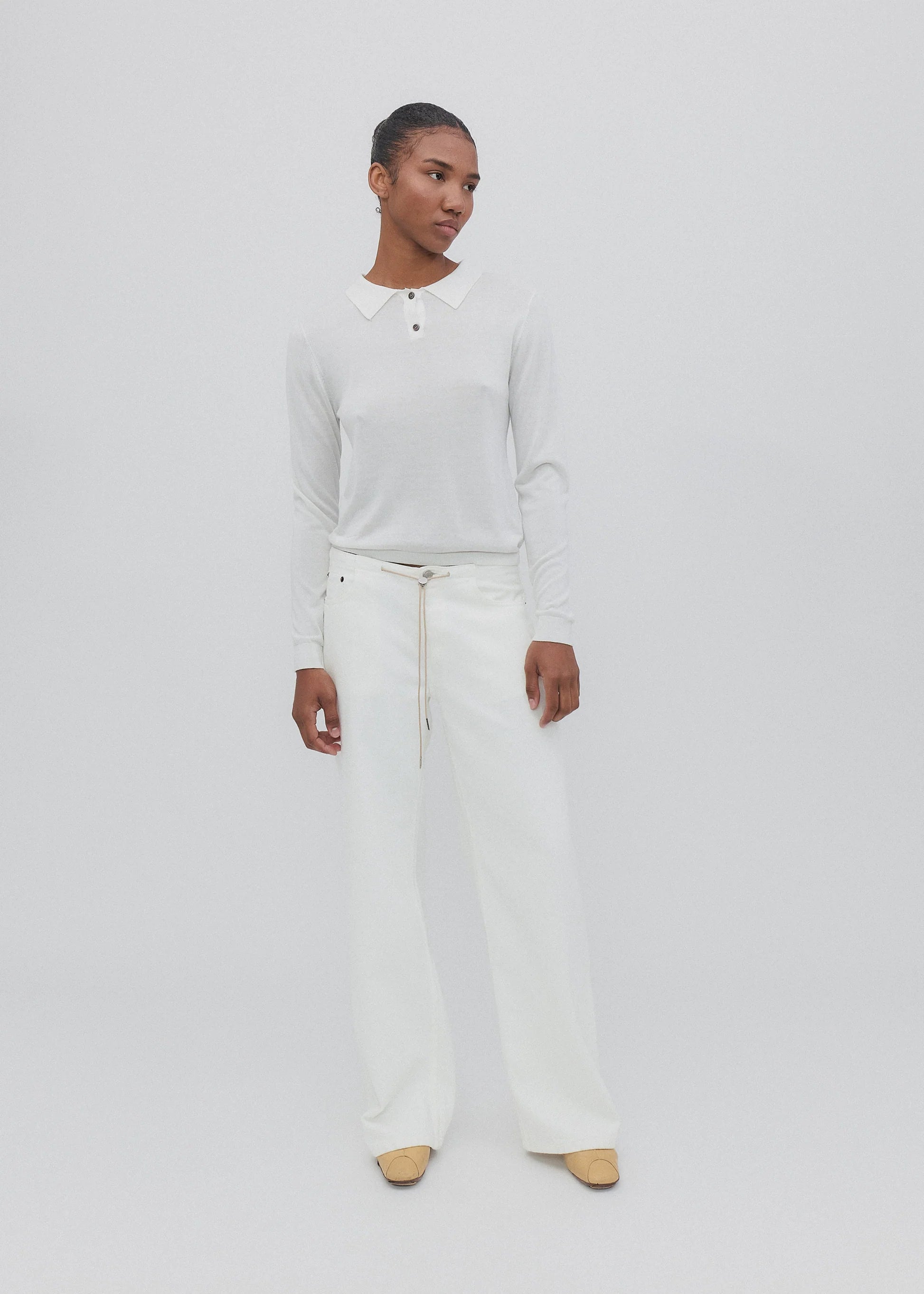 A person stands against a plain white background, wearing a white long-sleeved shirt with a collar, paired with the Cool Jean—white relaxed leg jeans featuring an adjustable drawstring—and beige shoes. They have short hair and a relaxed expression, with arms resting by their sides.