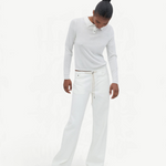 A person stands against a plain light background, looking downward with hands by their sides, wearing a white long-sleeve shirt and the Cool Jean—white relaxed leg jeans featuring an adjustable drawstring—paired with tan shoes.