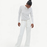 A person stands against a plain light background, looking downward with hands by their sides, wearing a white long-sleeve shirt and the Cool Jean—white relaxed leg jeans featuring an adjustable drawstring—paired with tan shoes.