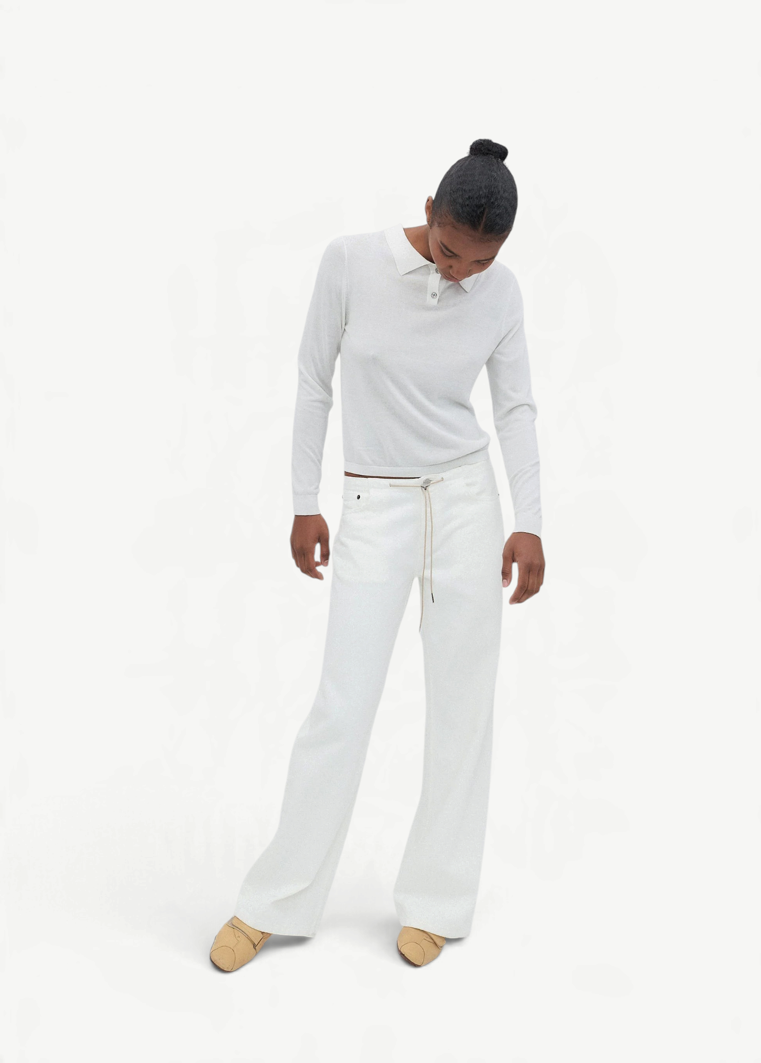 A person stands against a plain light background, looking downward with hands by their sides, wearing a white long-sleeve shirt and the Cool Jean—white relaxed leg jeans featuring an adjustable drawstring—paired with tan shoes.