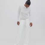 A person stands against a plain light background, looking downward with hands by their sides, wearing a white long-sleeve shirt and the Cool Jean—white relaxed leg jeans featuring an adjustable drawstring—paired with tan shoes.