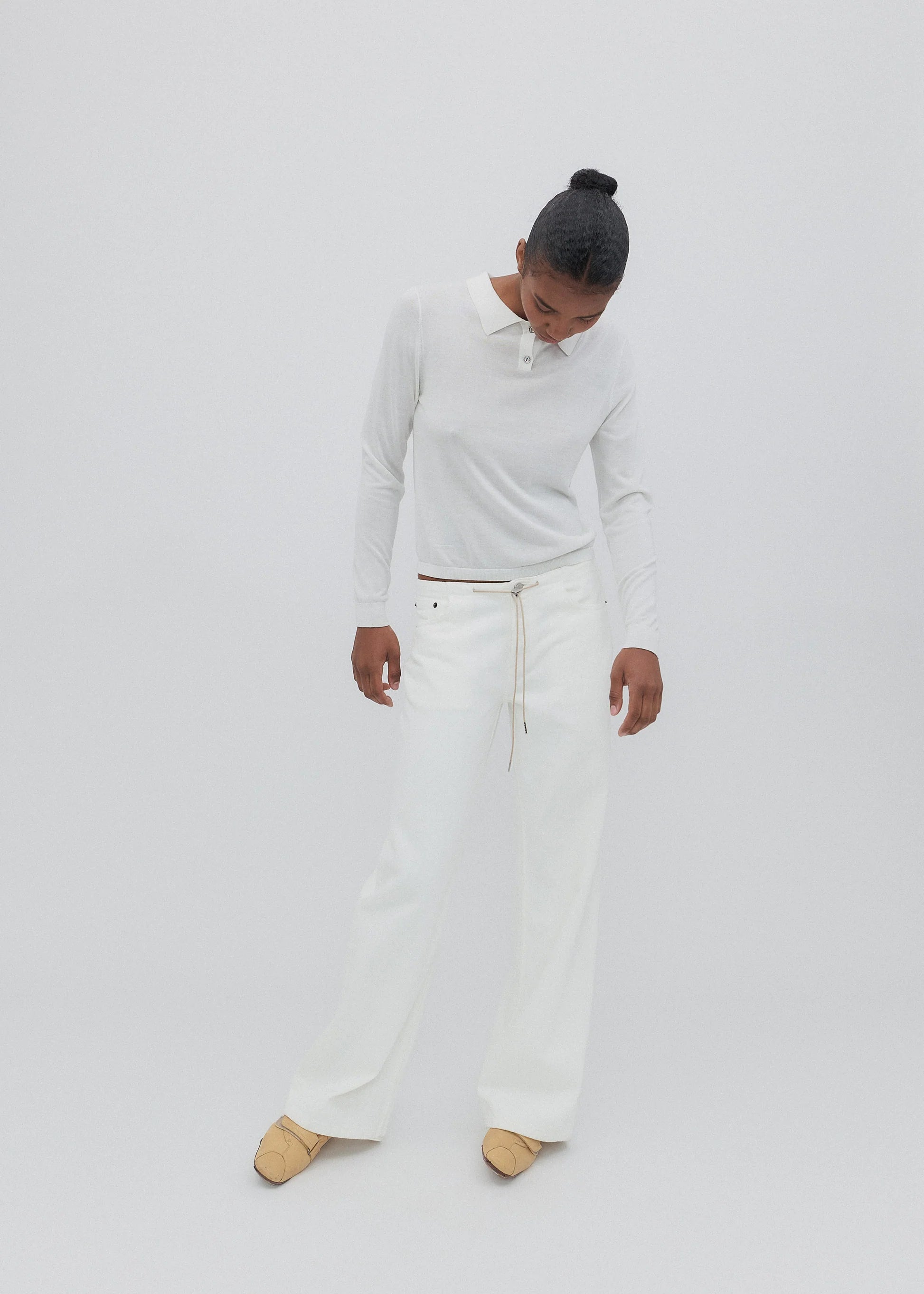 A person stands against a plain light background, looking downward with hands by their sides, wearing a white long-sleeve shirt and the Cool Jean—white relaxed leg jeans featuring an adjustable drawstring—paired with tan shoes.