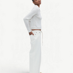 A person stands sideways against a plain white background, wearing a white long-sleeve top, tan shoes, and the Cool Jean—relaxed leg jeans featuring an adjustable drawstring.