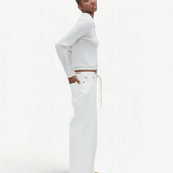 A person stands sideways against a plain white background, wearing a white long-sleeve top, tan shoes, and the Cool Jean—relaxed leg jeans featuring an adjustable drawstring.