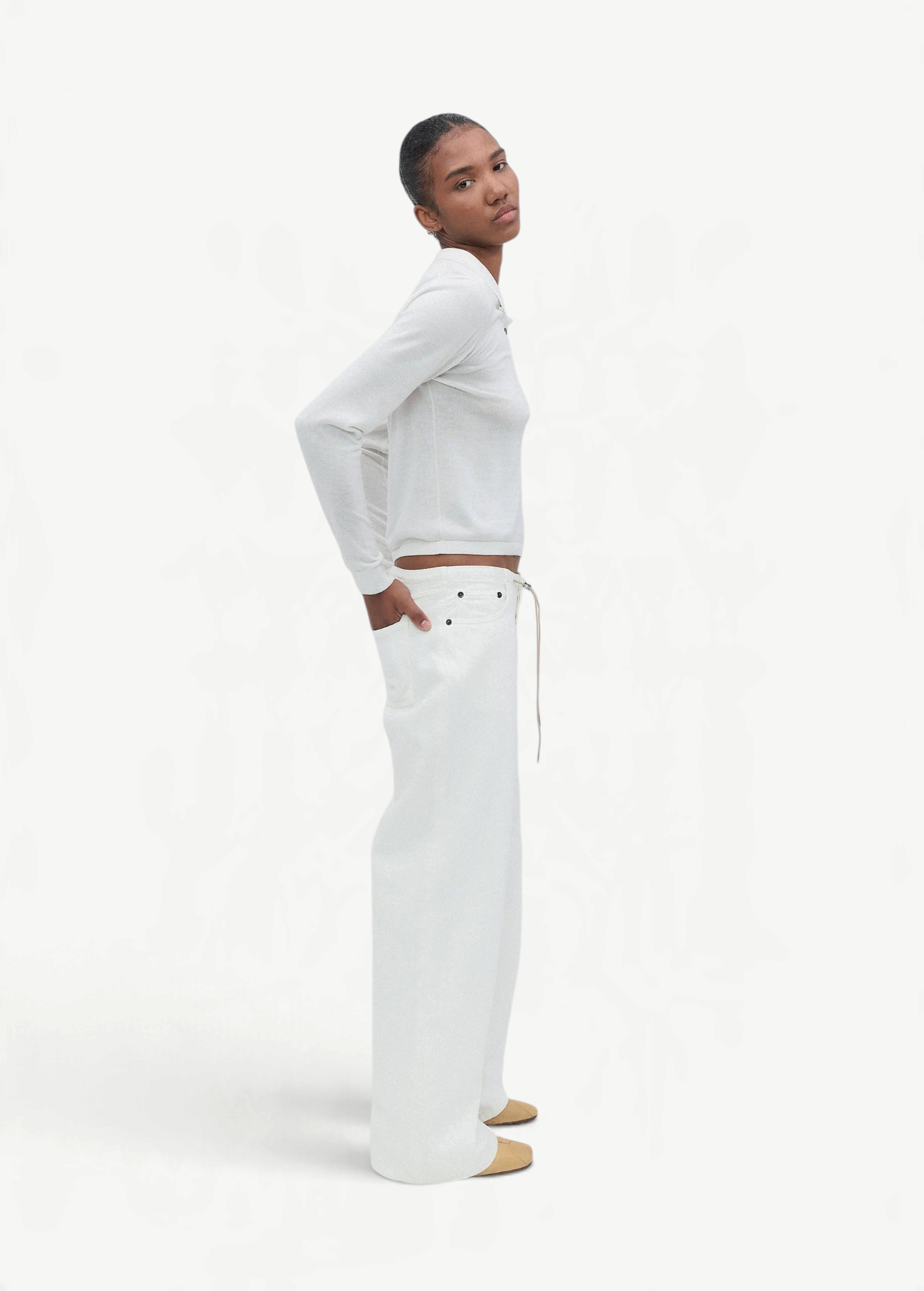 A person stands sideways against a plain white background, wearing a white long-sleeve top, tan shoes, and the Cool Jean—relaxed leg jeans featuring an adjustable drawstring.