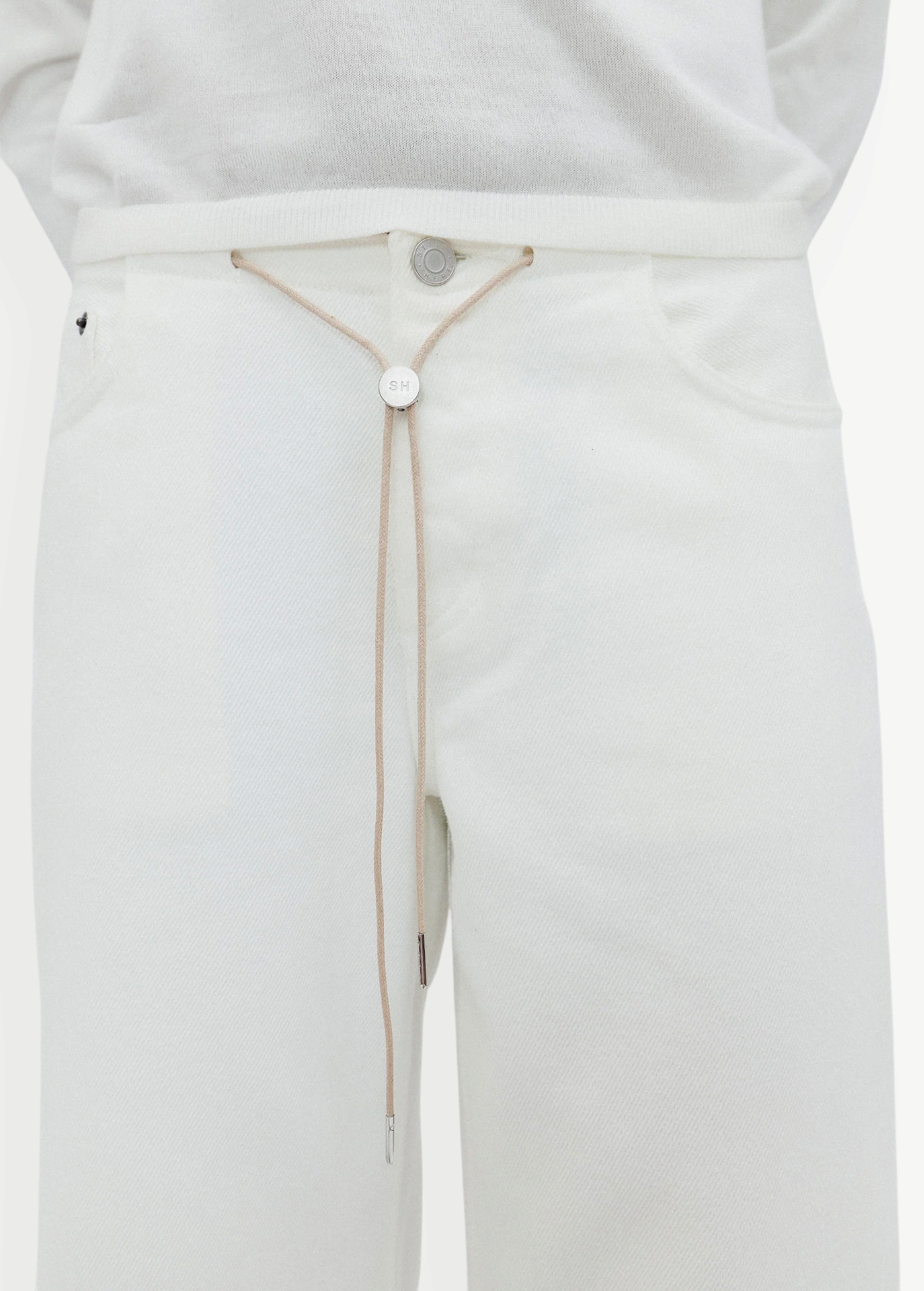 Wearing the Cool Jean in an off-white shade with a matching top, this look is cinched at the waist with a beige string belt featuring a round metallic front accent threaded through the waistband. The relaxed leg silhouette of the Cool Jean lends an effortlessly laid-back vibe to the outfit.