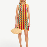 The Hazel Knit Dress is a sleeveless crochet mini dress featuring a colorful vertical zigzag pattern, styled with large circular earrings, tan thong sandals, and a raffia clutch.