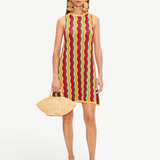 The Hazel Knit Dress is a sleeveless crochet mini dress featuring a colorful vertical zigzag pattern, styled with large circular earrings, tan thong sandals, and a raffia clutch.