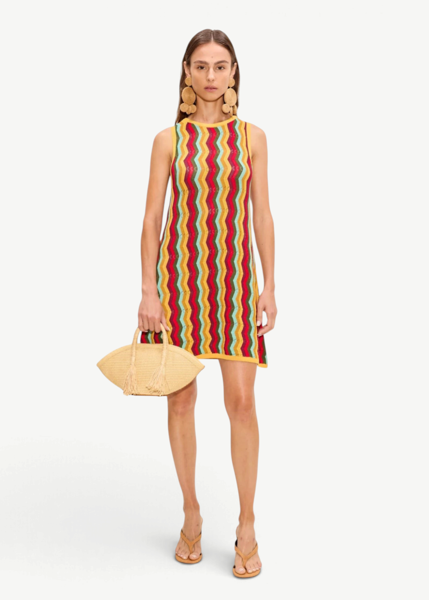 The Hazel Knit Dress is a sleeveless crochet mini dress featuring a colorful vertical zigzag pattern, styled with large circular earrings, tan thong sandals, and a raffia clutch.