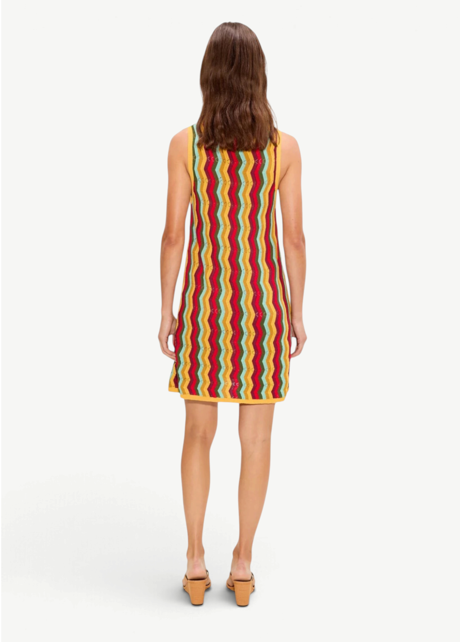 A person from behind wearing the Hazel Knit Dress, a sleeveless, knee-length crochet mini dress featuring a vertical multi-colored chevron pattern and yellow trim.