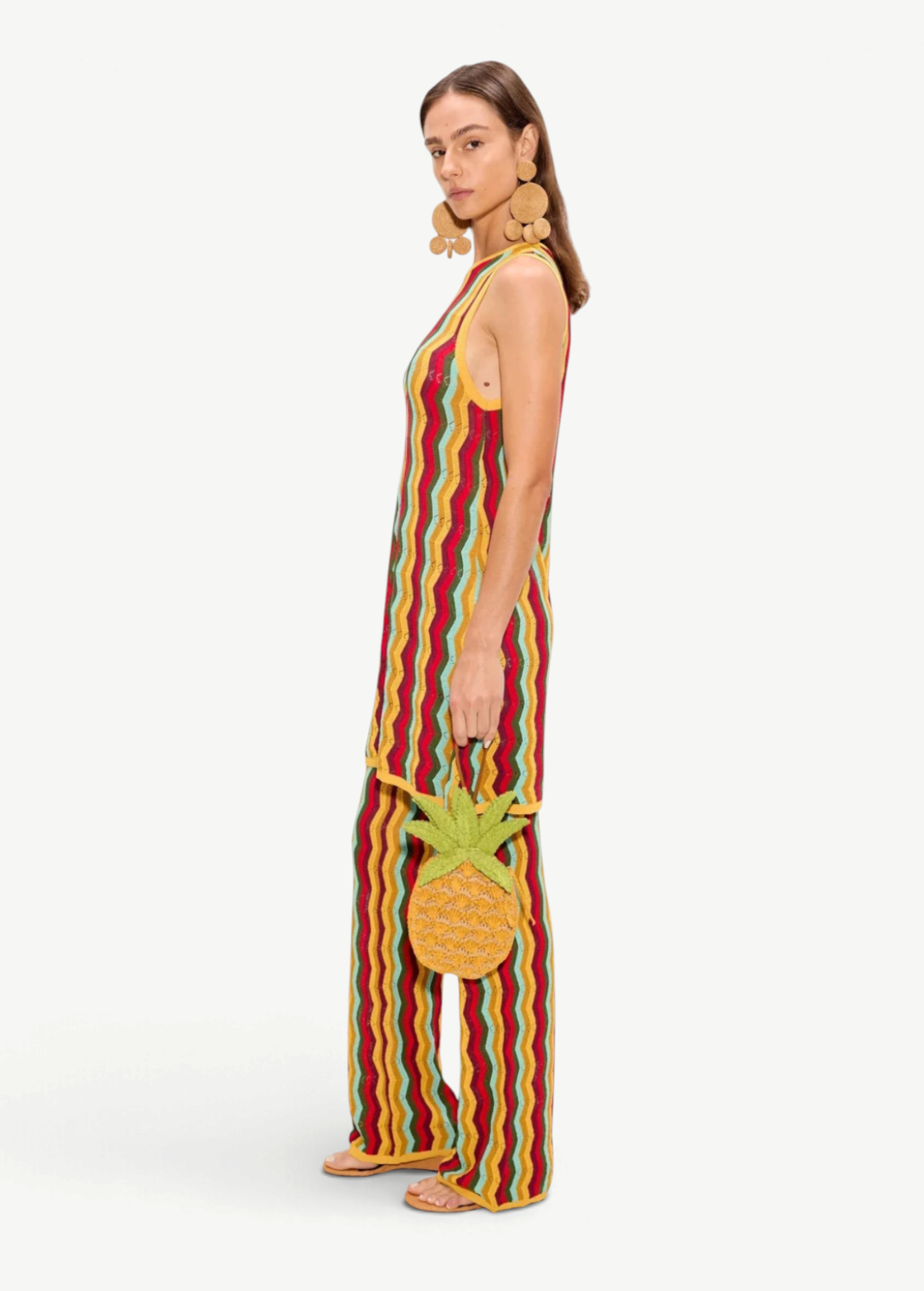 A person wears a Hazel Knit Dress, a sleeveless, floor-length crochet mini dress with a red, yellow, and green wavy pattern, carrying a small pineapple-shaped handbag.