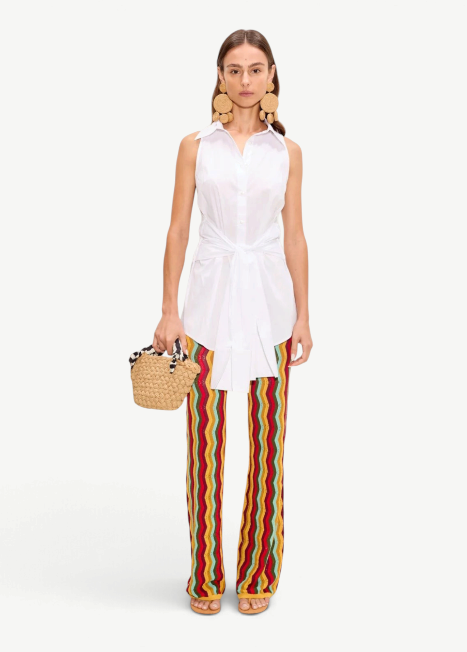 A woman wearing a white sleeveless button-down shirt tied at the waist and colorful chevron-striped Ophelia Knit Pant, carrying a small woven handbag.