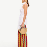 A person wearing a white sleeveless collared shirt and Ophelia Knit Pants featuring a colorful squiggle design, carrying a small woven handbag.
