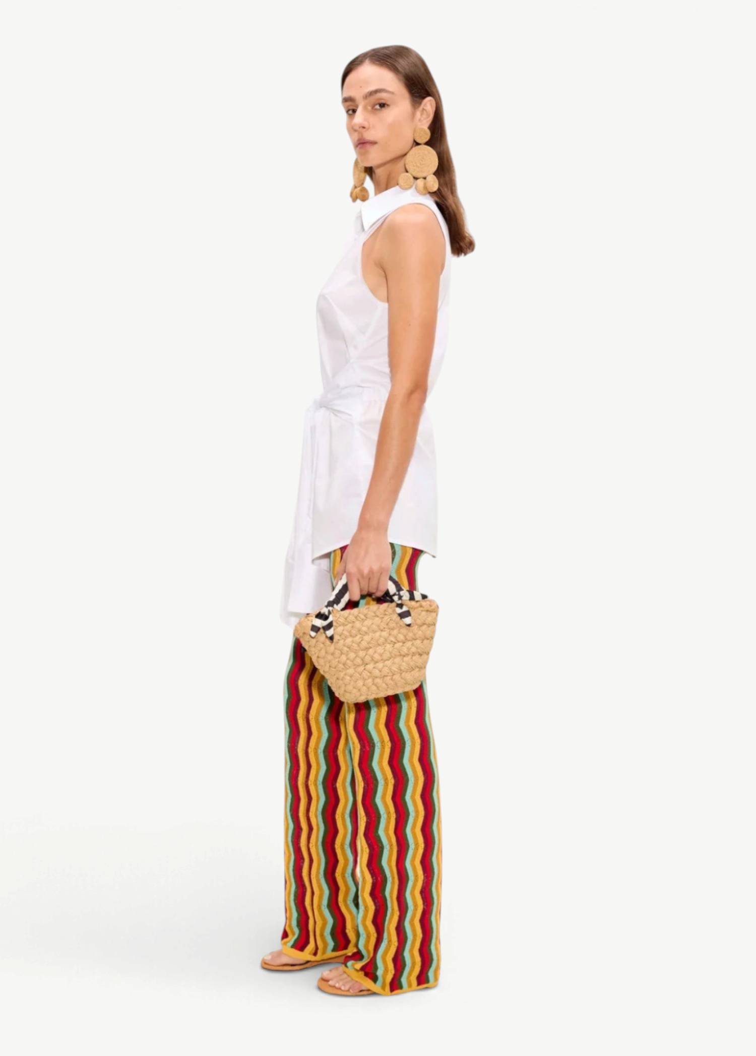 A person wearing a white sleeveless collared shirt and Ophelia Knit Pants featuring a colorful squiggle design, carrying a small woven handbag.