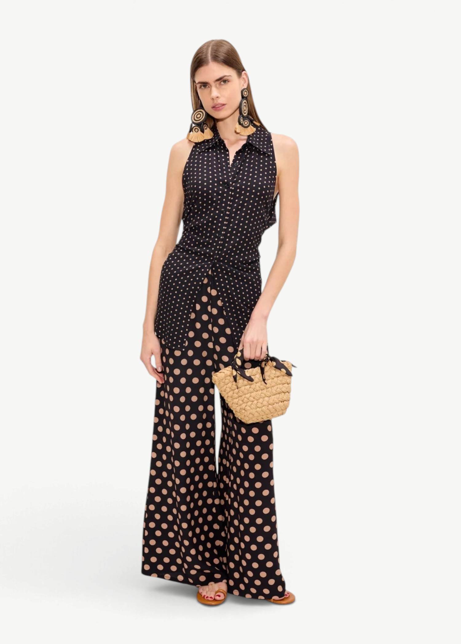 A model wears a black polka-dot Loch Sleeveless Crepe Button Up with matching wide-leg trousers and a small woven handbag.